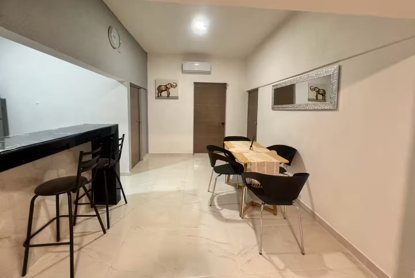 Une cuisine moderne avec un plan de travail noir et des tabourets de bar, un coin repas avec une table en bois et des chaises noires, et un couloir avec une porte et des œuvres d’art accrochées au mur.