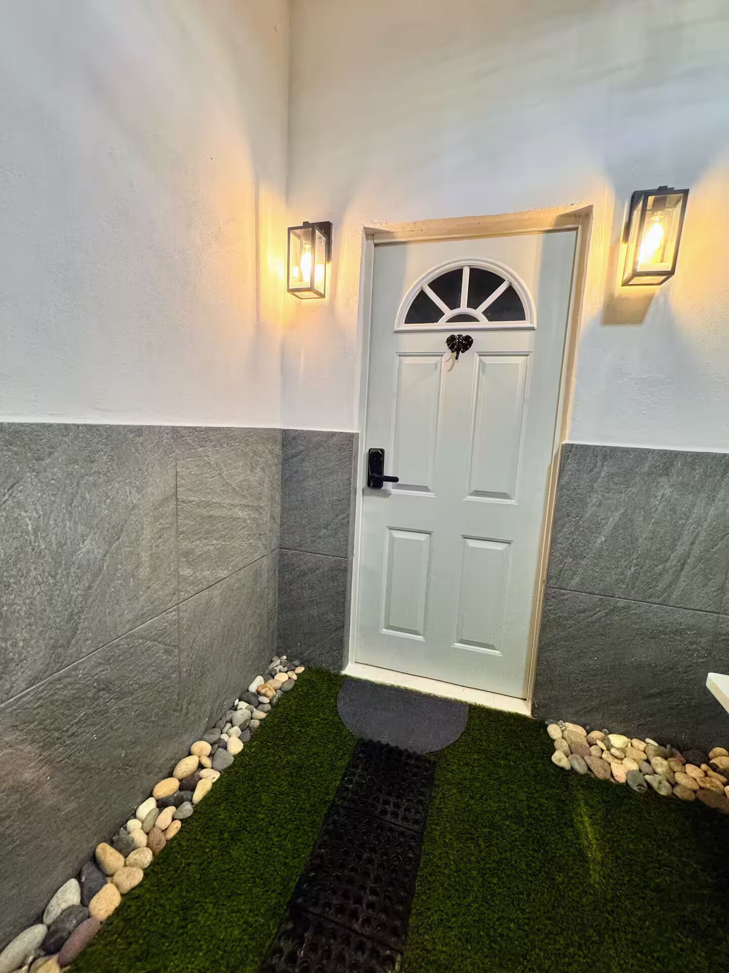 Une porte blanche ornée d’un motif décoratif en partie supérieure, flanquée de deux lampes fixées au mur, se trouve dans un couloir aux murs en pierre grise et dont la base est recouverte de petits galets.