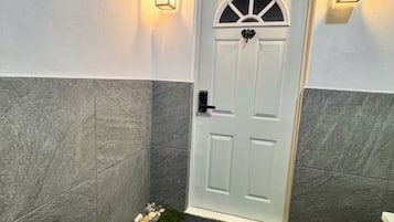 Une porte blanche ornée d’un motif décoratif en partie supérieure, flanquée de deux lampes fixées au mur, se trouve dans un couloir aux murs en pierre grise et dont la base est recouverte de petits galets.