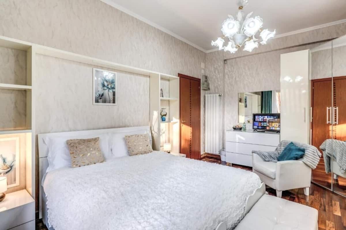 A bedroom with a large bed, a white chair, a dresser, and a mirror.