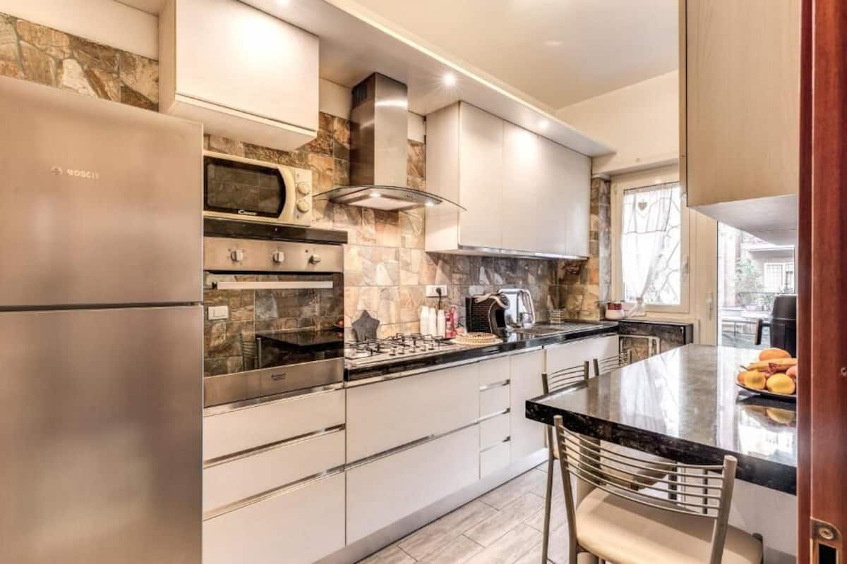 A modern kitchen with a stainless steel refrigerator, a built-in oven, a microwave, and a gas stove. There is a large island with a marble countertop and a chair.
