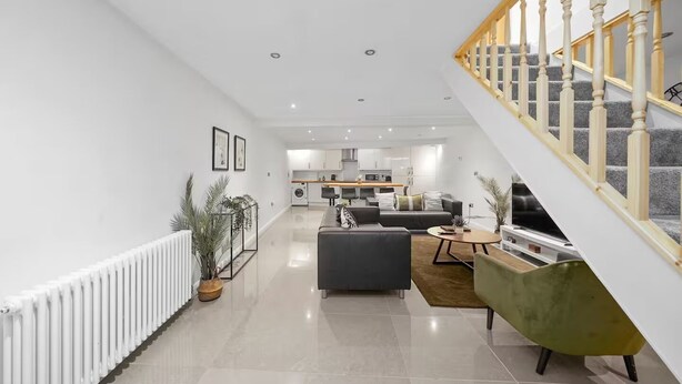 A modern living room with a staircase, a kitchen in the background, and a radiator on the left. - Fully Equipped 2BR Flat | Family|Relocations| ST (Greater London)