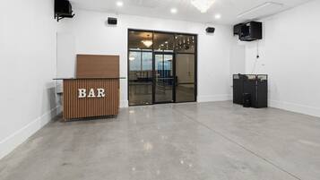 A modern bar area with a wooden counter labeled 'BAR', a glass door leading outside, and a black speaker system on the wall.