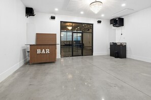 Un área de bar moderna con mostrador de madera identificado con la leyenda 'BAR', una puerta de vidrio que da al exterior y un sistema de altavoces negros en la pared.