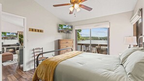 Une chambre avec un lit, un ventilateur de plafond, une commode, une chaise, un téléviseur et une vue sur un lac, visible à travers de grandes fenêtres.