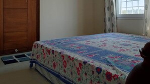 Une chambre avec une armoire en bois, un lit recouvert d’une couvre-lit à motifs floraux, une fenêtre avec des rideaux et un petit tapis sur le sol.