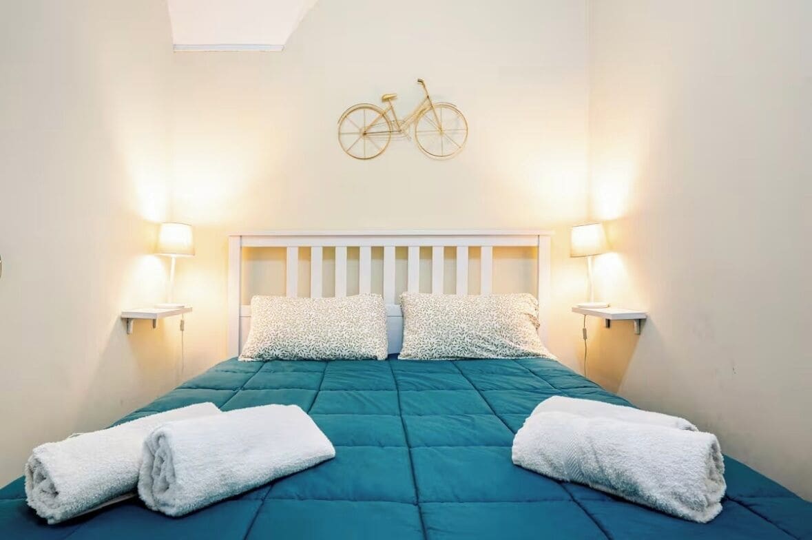 Un lit bien fait, avec une couette turquoise, des oreillers blancs et deux serviettes enroulées posées sur le côté.