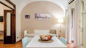 Une pièce confortable avec un lit, une table à manger agrémentée d’un panier de fruits, une lampe et un tableau accroché au mur.