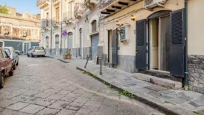 Une ruelle étroite pavée, bordée de bâtiments historiques, dont certains ont leurs volets fermés, et où des voitures sont garées le long du trottoir.