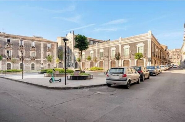 A street with parked cars and a pedestrian area with a bench. - Spàzio apartments Archi (Catania)