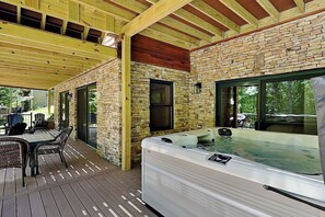 Un espacio al aire libre techado con un jacuzzi, terraza de madera, paredes de piedra y una mesa de patio con sillas.