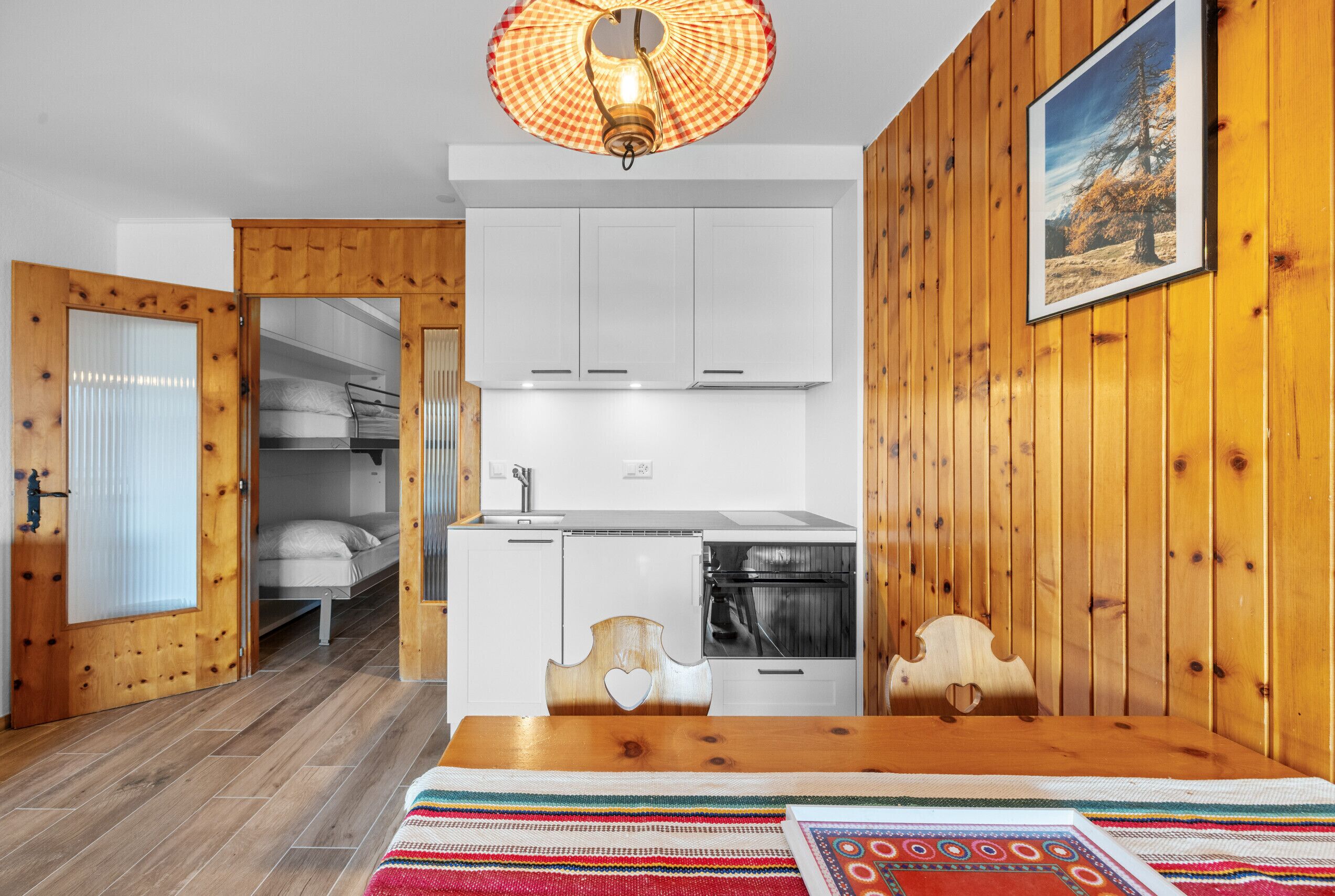 A compact room with a wooden floor, a bed with a white headboard, a kitchenette with white cabinets, a stove, and a hanging light fixture.