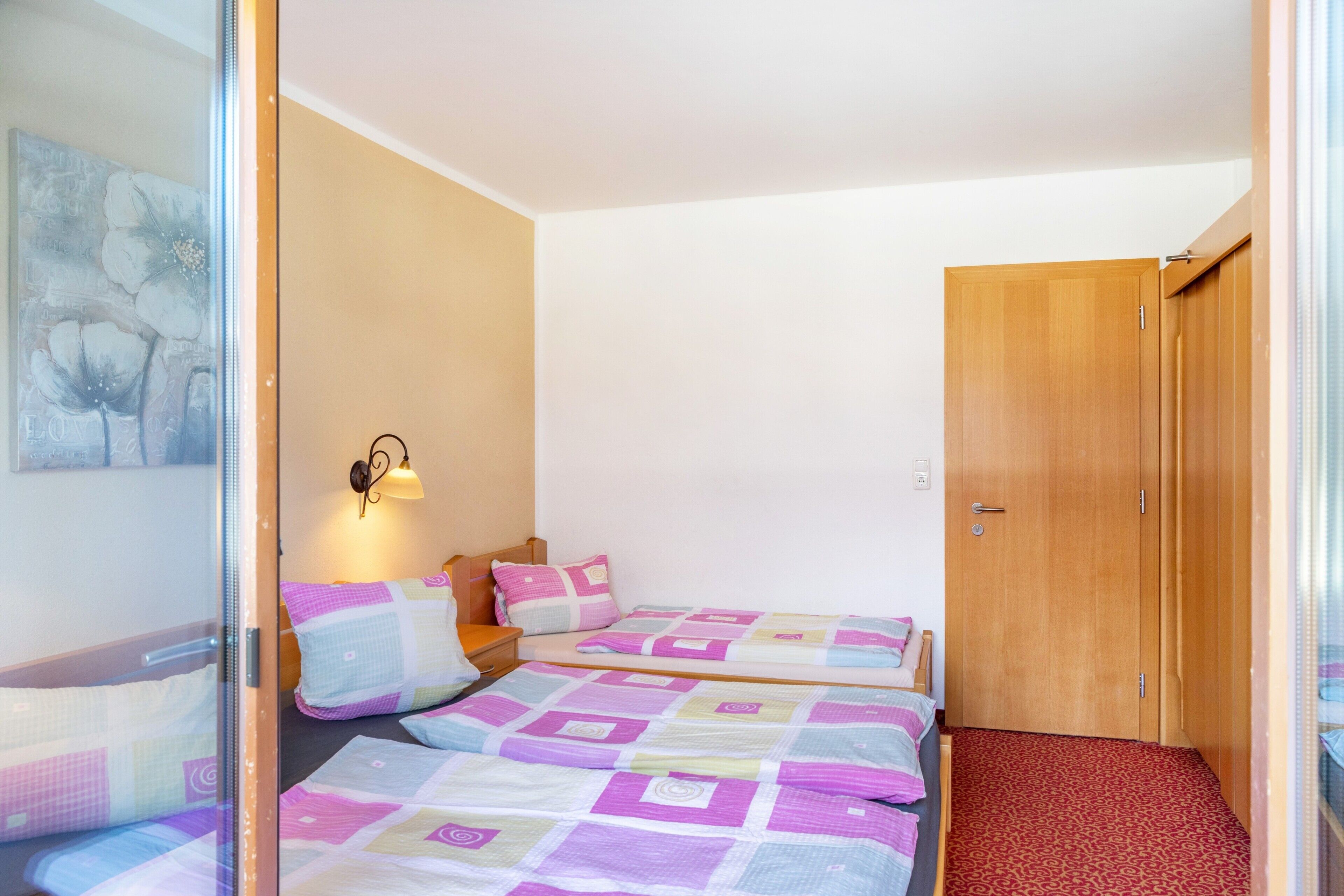 A bedroom with two beds, a wooden wardrobe, a wall-mounted lamp, and a painting.