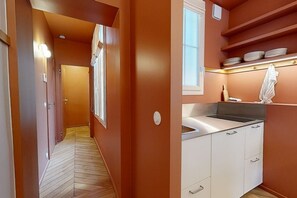Un couloir étroit, doté d’un plancher en bois, mène à une cuisine avec des meubles blancs et un évier.