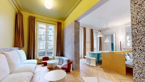 A modern living room with a sofa, coffee table, and chairs, a small kitchen area, and a view of the city through large windows. - Joivy Delightful 1-bed Studio w/ Shared Co-working (Paris)