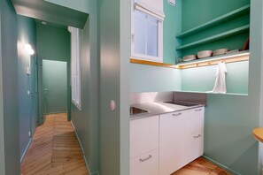 Un couloir étroit doté d’un parquet en bois disposé en chevrons mène à une cuisine avec des meubles blancs, un évier en acier inoxydable et une fenêtre équipée de stores.