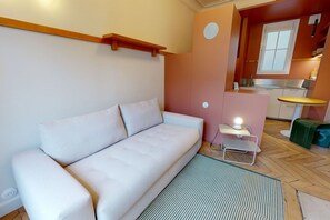 Un salon moderne avec un canapé blanc, une petite table d’appoint avec une lampe, et un coin cuisine à l’arrière-plan.