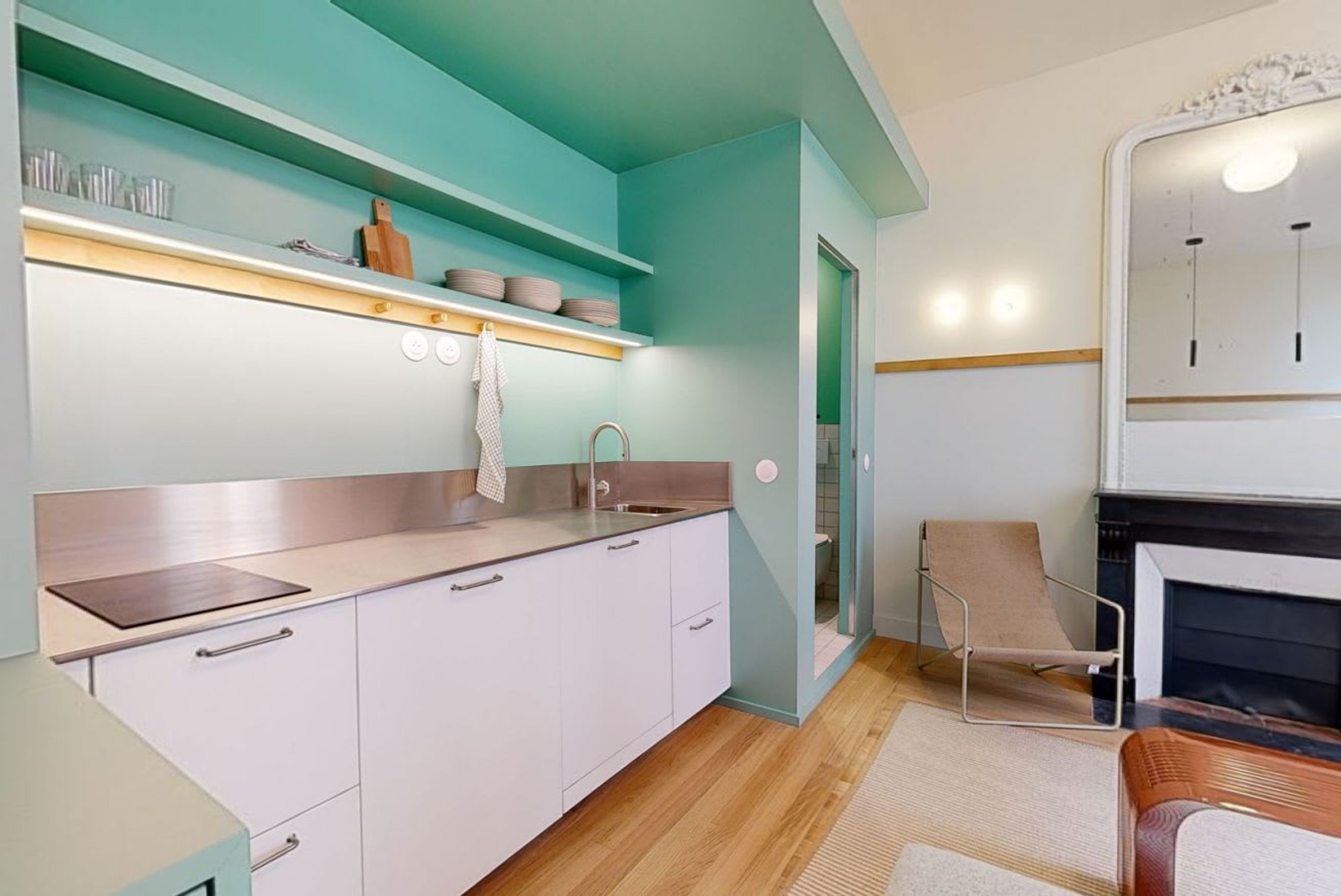 A modern kitchen with white cabinets, a stainless steel countertop, and a teal backsplash. There is a wooden chair and a fireplace in the adjacent living area.