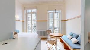 Una camera d'albergo con un letto, un tavolino rotondo, una poltrona e ampie finestre che danno su edifici.