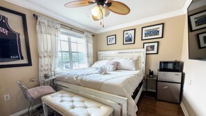 A bedroom with a bed, a bench, a chair, a ceiling fan, a window with curtains, a refrigerator, and framed pictures on the wall. - Home located  10 minute from Hard Rock Stadium Home of the FIFA World Cup 2026  (Miramar)