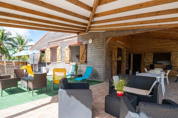 Un patio cubierto al aire libre con mobiliario de mimbre, una mesa de comedor y una cocina visible a través de una puerta abierta.