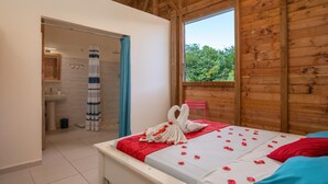 Ein Schlafzimmer mit Holzbalkendecke, einem Bett mit rot-blauer Bettwäsche, einem Fenster mit Blick auf Bäume und einem Badezimmer mit Duschvorhang.