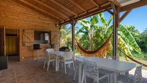 Eine Holzblockhütte mit einer gefliesten Terrasse, weißen Möbeln und einer Hängematte mit Blick auf üppiges Grün.