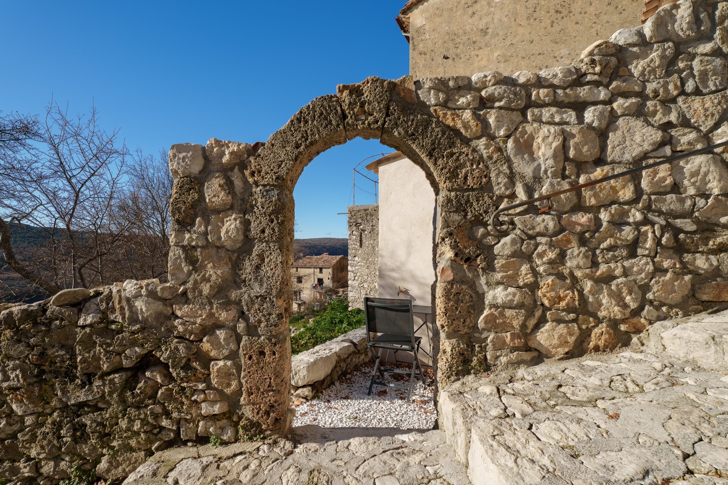 An ancient stone archway leading to a courtyard with a table and chair, surrounded by stone walls and a clear sky.