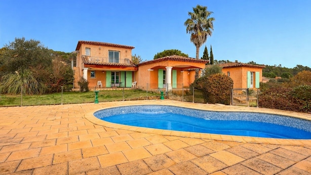 Una casa de dos pisos con piscina, rodeada de vegetación y un cielo azul despejado.