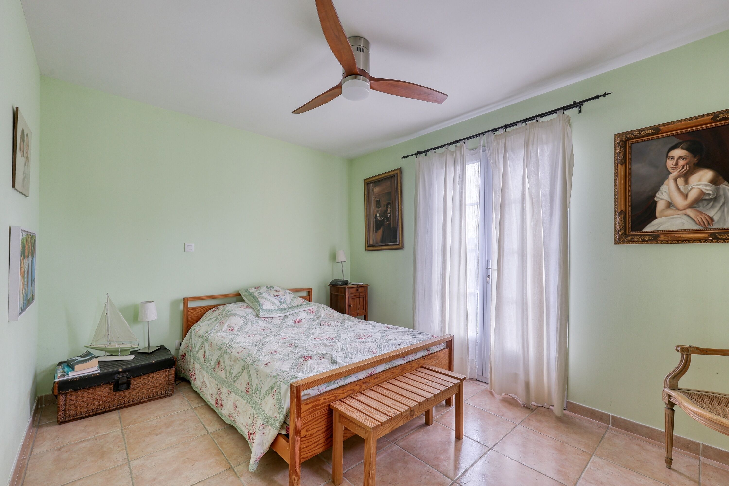 A bedroom with a wooden bed, a bench, a suitcase, a ceiling fan, and a painting on the wall.