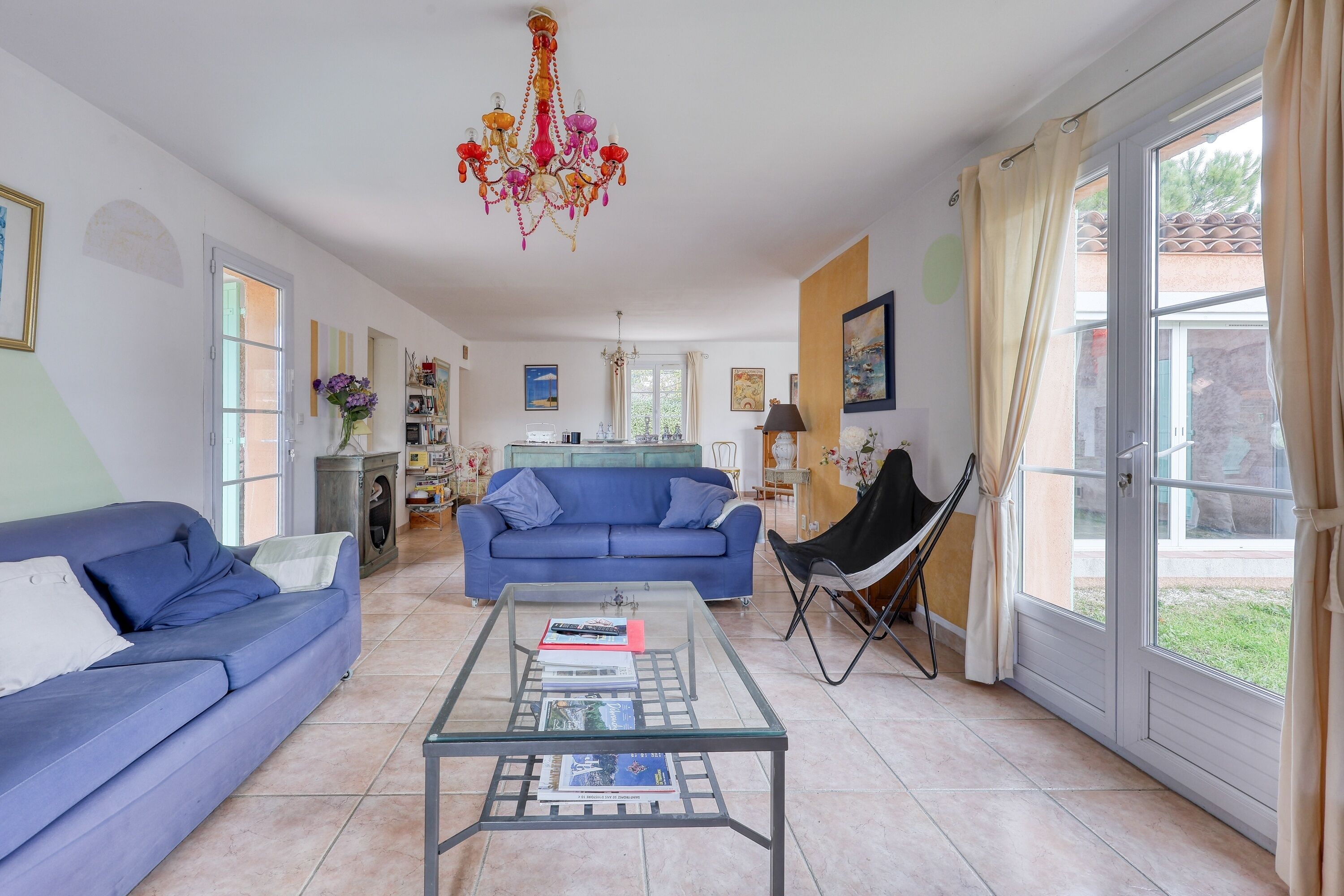 Una sala de estar con un sofá azul, una mesa de centro de vidrio con libros, una araña y vistas a través de un amplio ventanal.