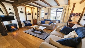 A cozy living room with a fireplace, sofas, a television, and a window with curtains. - Honeycott | Sleeps 4 | Croyde Beach (Croyde)