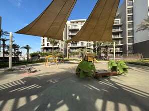 Un parque infantil con columpios y un balancín, rodeado de palmeras y edificios.