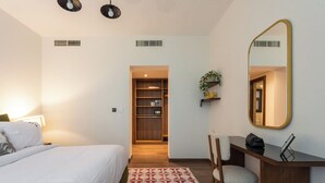 A hotel room with a bed, a desk, a chair, a mirror, a shelf with a plant, and a patterned rug. - Chic 3BR Garden View Apart at Cairo Festival City (Cairo Governorate)