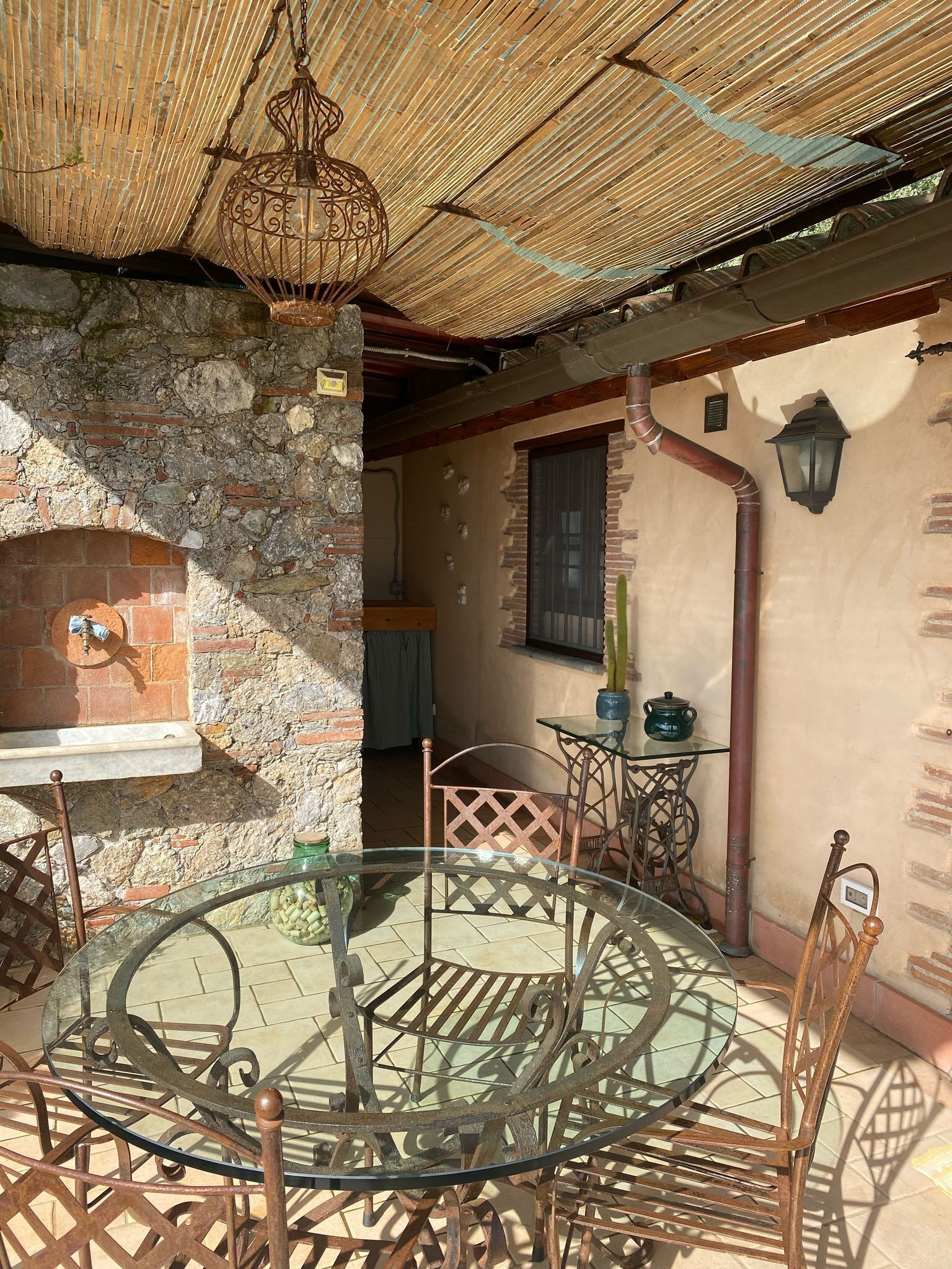 Une terrasse extérieure rustique avec une table ronde en verre et des chaises en métal, un four en briques et une lanterne suspendue.