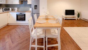 Una cocina moderna con armarios blancos, un microondas y una estufa. Hay un comedor con una mesa de madera y sillas blancas. Un televisor de pantalla plana está sobre un mueble apoyado contra la pared.