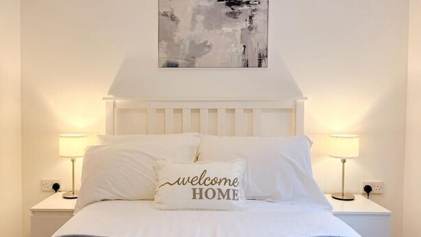 Una cama bien hecha con un cabecero blanco, dos lámparas de noche y una almohada con la inscripción “welcome HOME”.