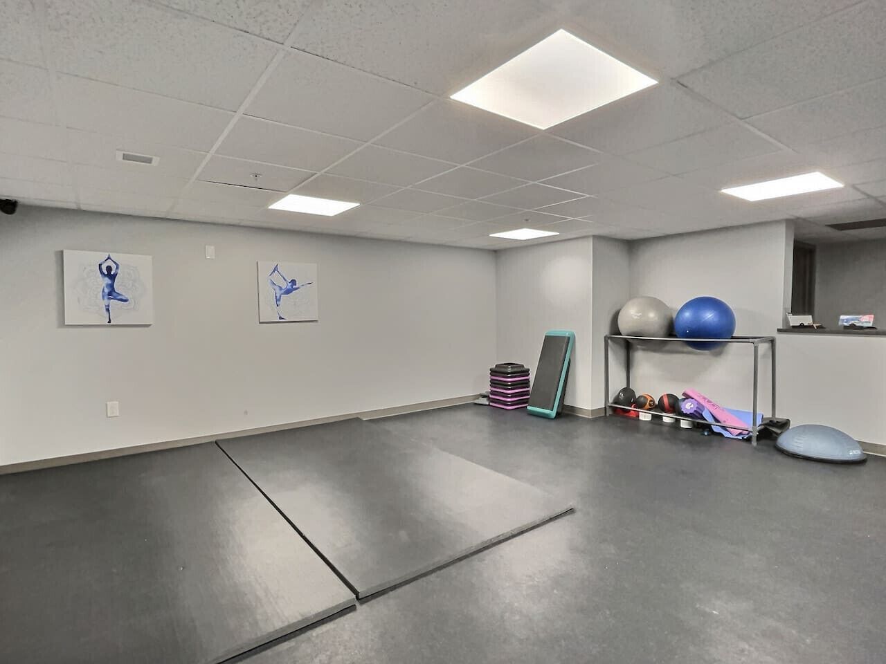 Une pièce spacieuse avec un plancher gris, des murs blancs et des luminaires au plafond. On y trouve du matériel d’exercice, comme une planche d’équilibre, un tapis de yoga et des ballons d’exercice.