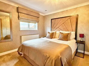 A bedroom with a large bed, a window with blinds, a radiator, a mirror, and a bedside table with a lamp. - Charming 3 bedroom house in Chelmsford (Essex)