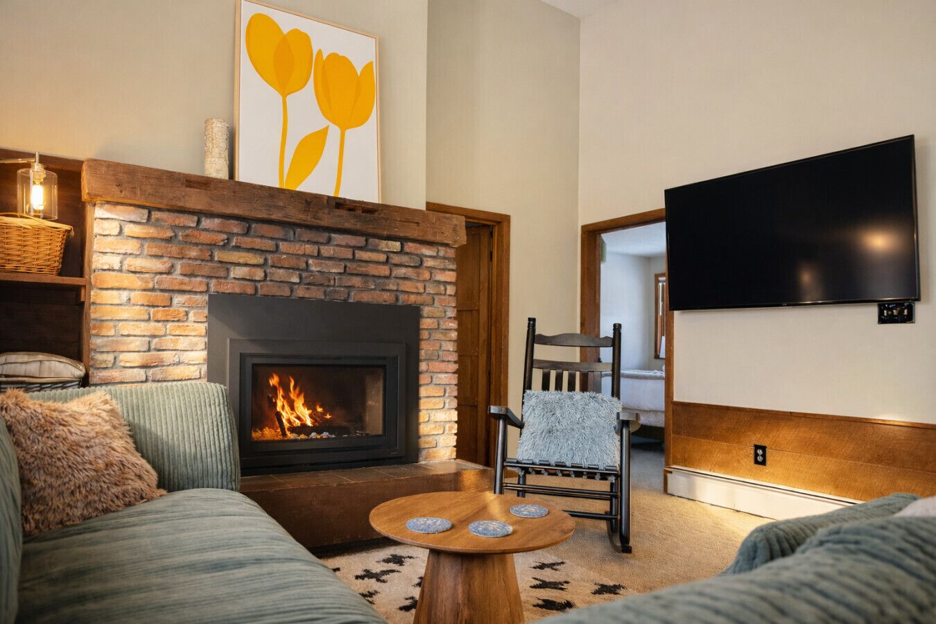 A living room with a brick fireplace, a sofa, a chair, a coffee table, and a flat-screen TV mounted on the wall.
