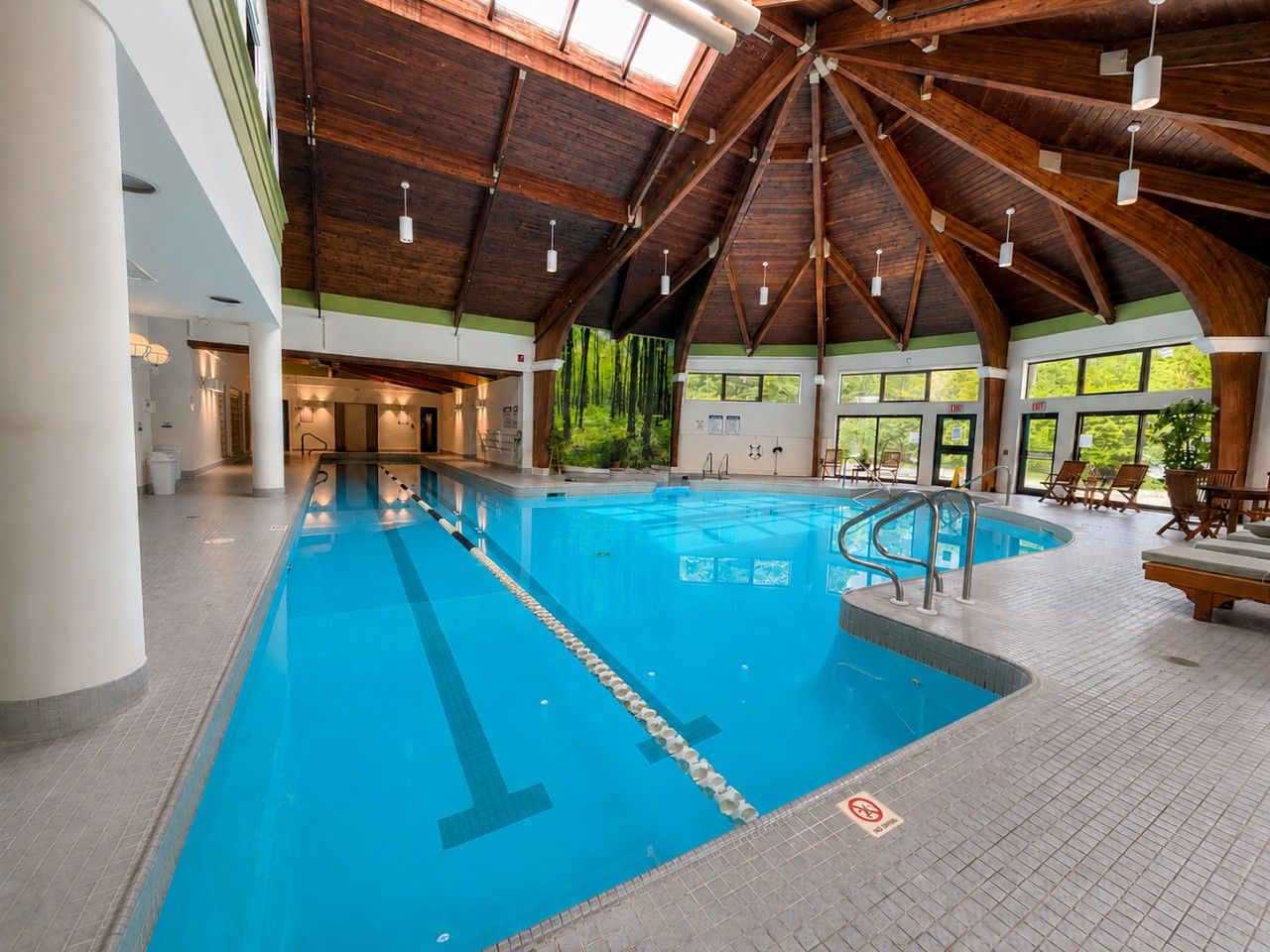 An indoor swimming pool with a wooden ceiling and tiled floor, surrounded by lounge chairs and a view of greenery outside.