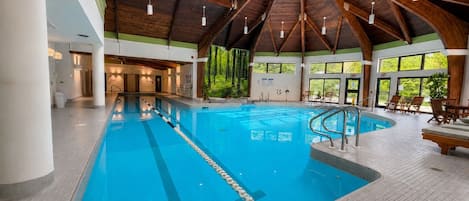 An indoor swimming pool with a wooden ceiling and tiled floor, surrounded by lounge chairs and a view of greenery outside.