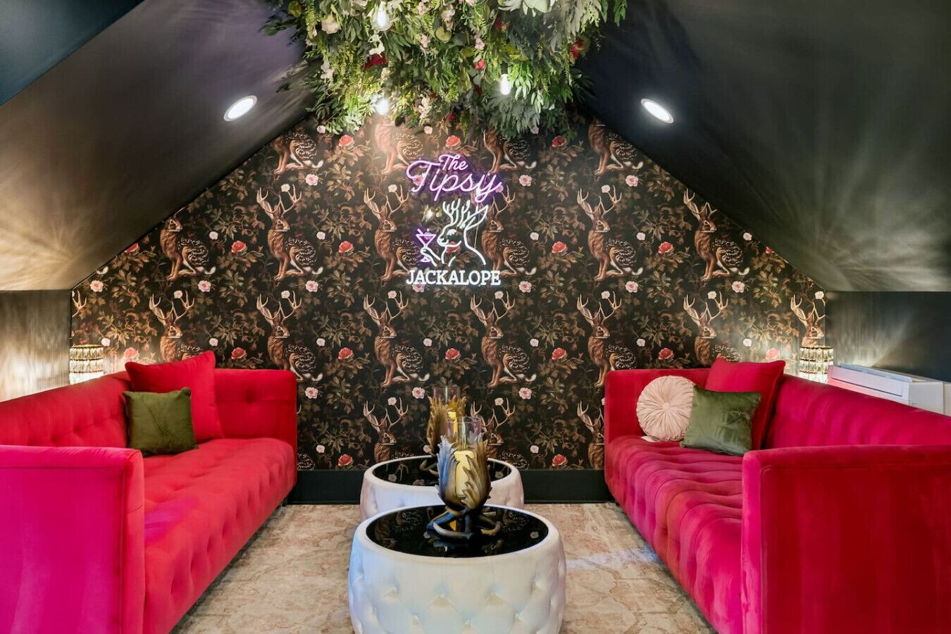 Ein gemütlicher Raum mit zwei roten Sofas, einer Wand mit Hirsch- und Blumenmuster sowie einem Neonlichtschild mit der Aufschrift "The Tips JACKALOPE".