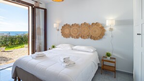 Une chambre avec un lit, deux lampes fixées au mur, une petite table avec une plante, et une vue sur la mer par une porte vitrée.