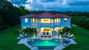 Una casa de dos pisos con piscina, rodeada de césped cuidado y palmeras.