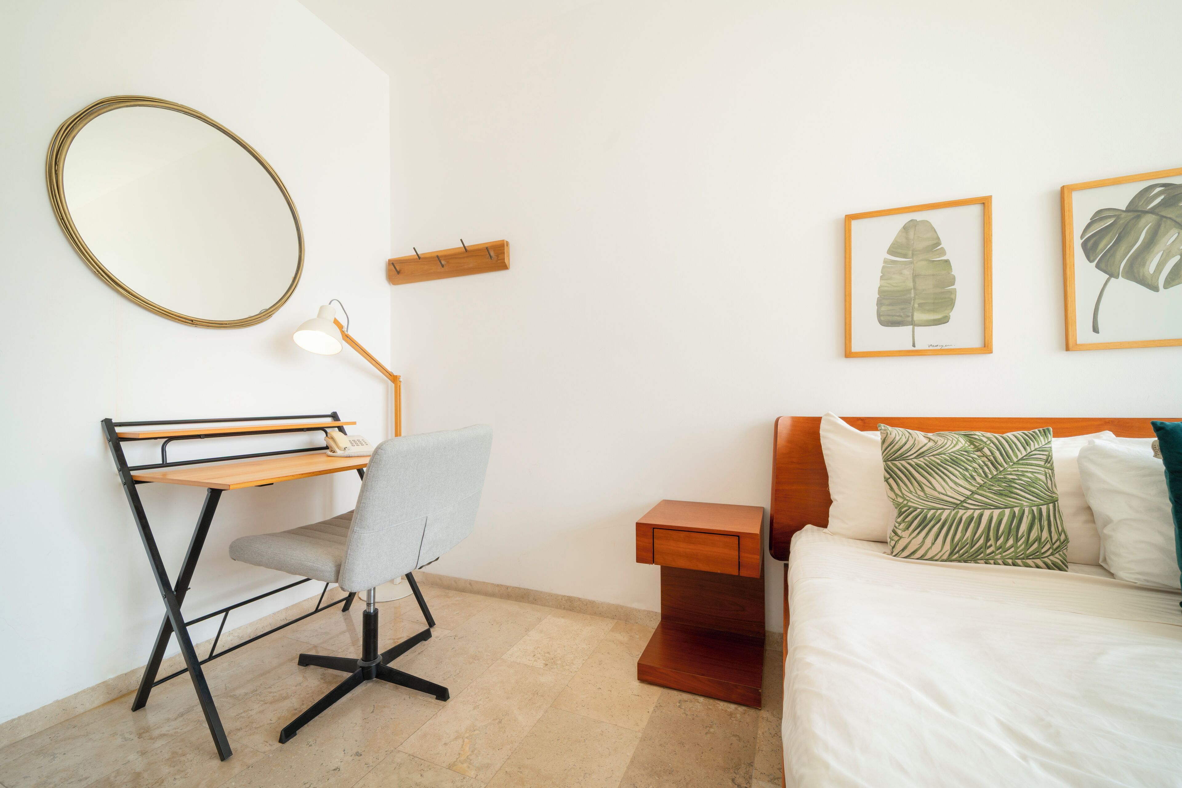 A bedroom with a bed, a desk with a chair, a round mirror, and two framed leaf artworks on the wall.