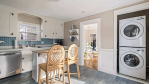 A modern kitchen with white cabinetry, a built-in dishwasher, a double washing machine, and a dining area with wicker chairs. - Designer Cottage • Game Room w/PacMan • Mins to Beach (Kennebunk)