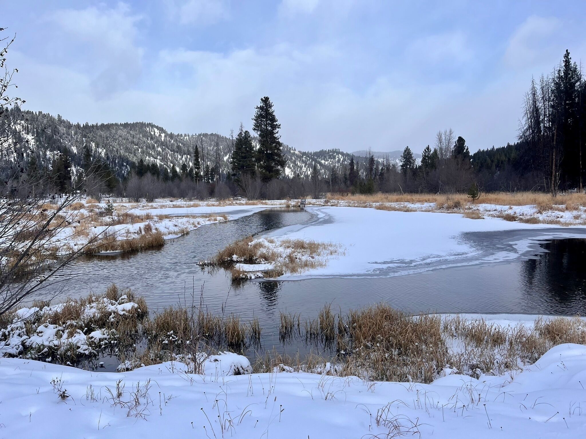 A winter landscape with a frozen river, snow-covered ground, and a forested mountain in the background.