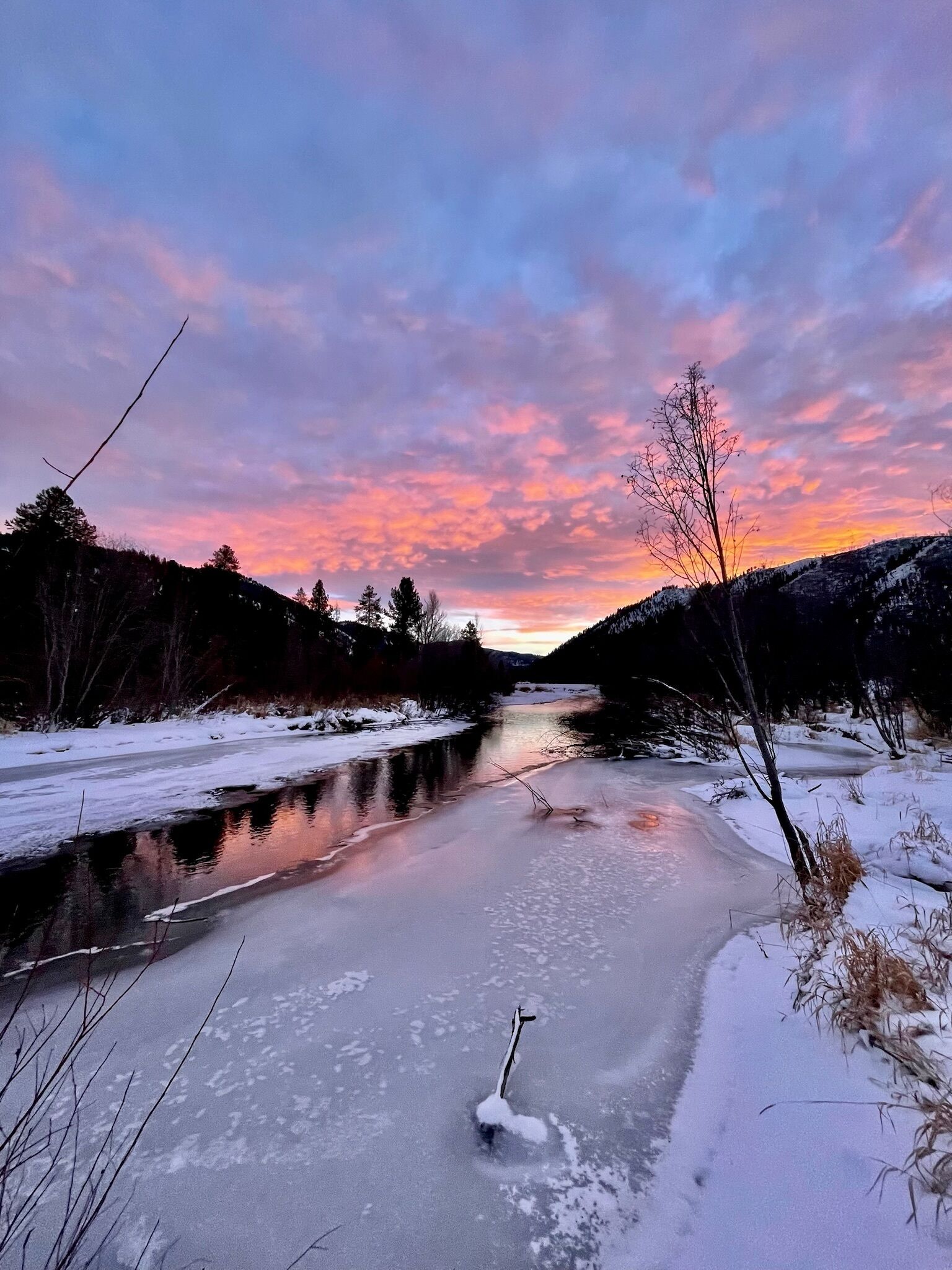 Une rivière gelée, avec un ciel aux couleurs du coucher de soleil, entourée de collines enneigées et de quelques arbres clairsemés.