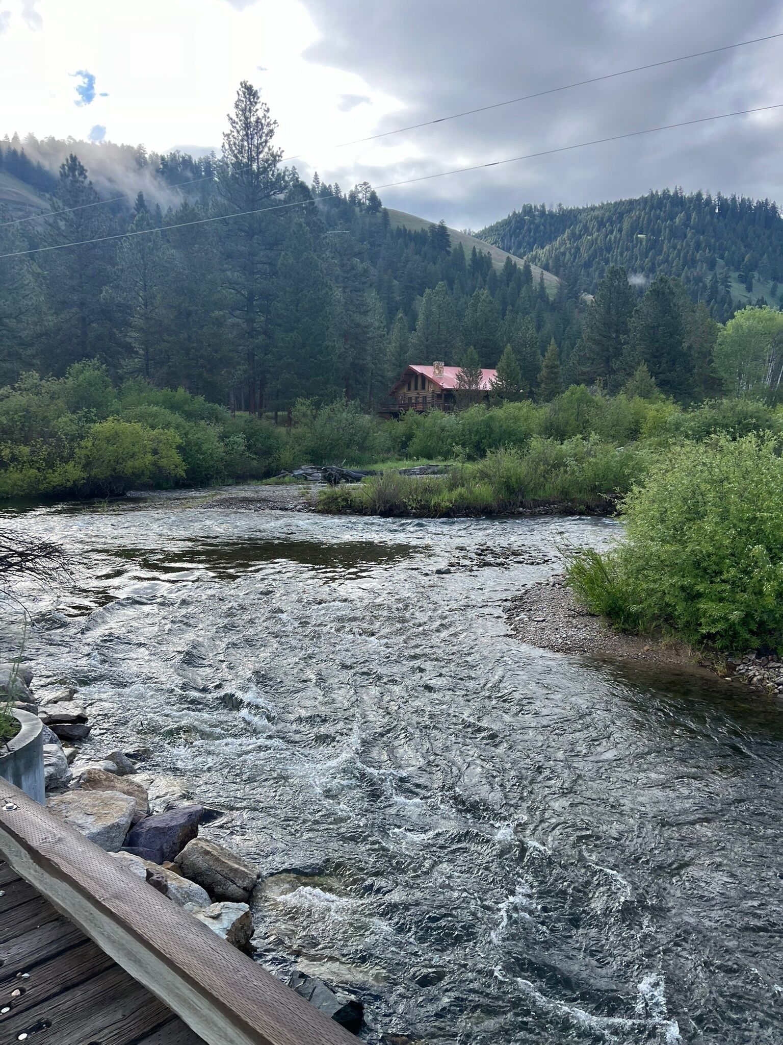 A river with a wooden bridge in the foreground, surrounded by dense forest and a mountain in the background.